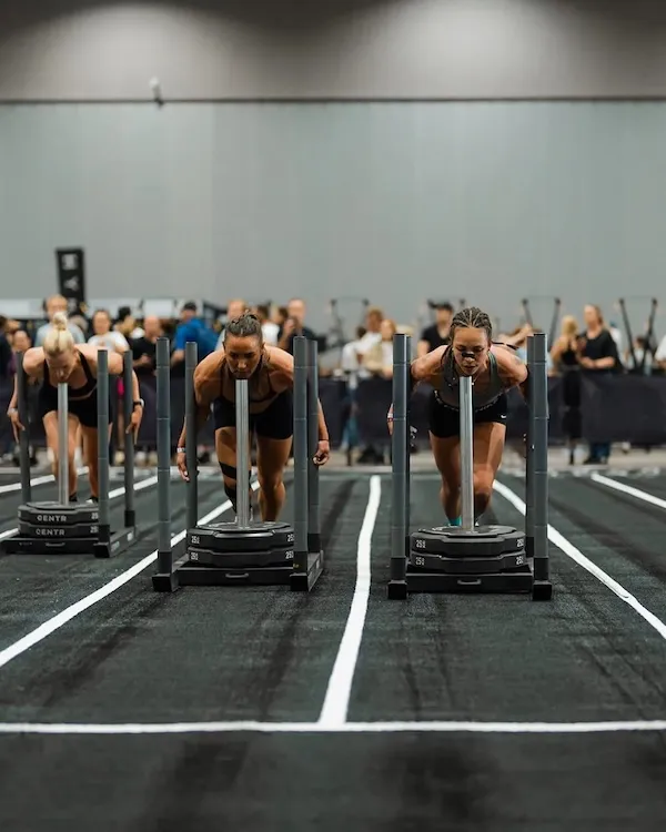 Three athletes pushing weighted sleds on parallel indoor lanes.